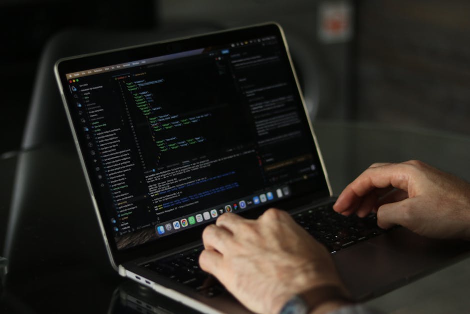 A close-up view of a person's hands typing on a laptop computer placed on a table, with the screen displaying lines of source code in a dark-themed text editor. The individual is working in a well-lit